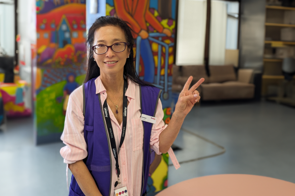 Austin Health volunteer Donna Ng wearing her purple volunteer vest, smiling at the camera and holding up the 'peace' symbol